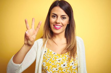 Genç güzel kadın sarı izole arka plan üzerinde duran ceket giyen gösteren ve emin ve mutlu gülümserken parmakları ile üç numara işaret.