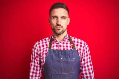 Genç yakışıklı adam yüzünde ciddi bir ifade ile kırmızı izole arka plan üzerinde dükkan sahibi önlük üniforma giyiyor. Kameraya basit ve doğal bir bakış.