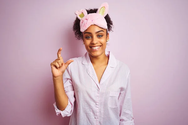 年轻的非洲裔美国妇女穿着睡衣和面具在孤立的粉红色背景微笑和自信的手势与手做小尺寸的标志用手指看和相机 测量概念 — 图库照片