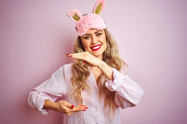 Young beautiful woman wearing pajama and sleep mask over pink isolated background gesturing with hands showing big and large size sign, measure symbol. Smiling looking at the camera. Measuring concept.