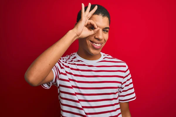 年轻的英俊阿拉伯男子穿着条纹T恤在孤立的红色背景做Ok手势与手微笑 眼睛透过手指看与快乐的脸 — 图库照片