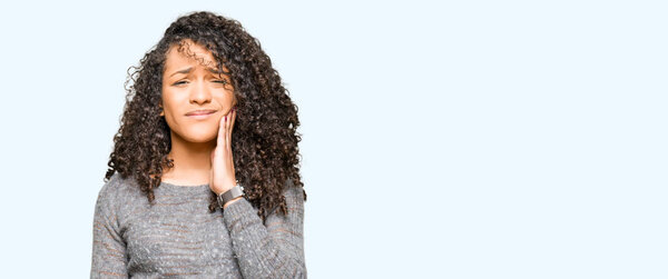 Young beautiful woman with curly hair wearing grey sweater touching mouth with hand with painful expression because of toothache or dental illness on teeth. Dentist concept.