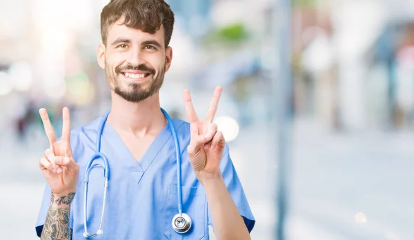 年轻英俊的护士男子穿着外科医生制服在孤立的背景微笑着看着镜头显示手指做胜利的标志 — 图库照片