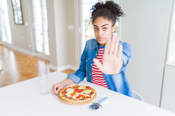 Young african american woman eating homemade mozzarella cheese pizza with open hand doing stop sign with serious and confident expression, defense gesture