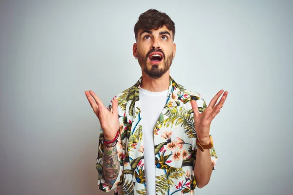 Tatilde dövmeli adam yazlık gömlek giyen izole beyaz arka plan üzerinde duran deli ve deli bağırarak ve agresif ifade ve kollar kaldırdı ile bağırıyor. Hayal kırıklığı kavramı.