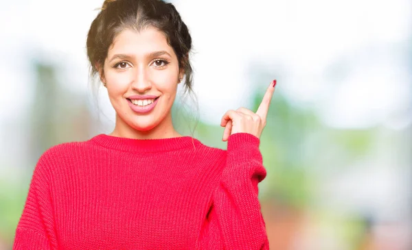 赤いセーターとパンを着た若い美しい女性は カメラを見て横に手と指で指差し 顔に大きな笑顔を浮かべて — ストック写真
