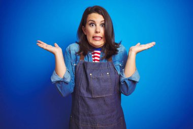 Genç güzel iş kadını mavi izole arka plan üzerinde mağaza üniforma önlük giyen habersiz ve kollar ve eller kaldırdı ile karışık ifade. Şüphe kavramı.