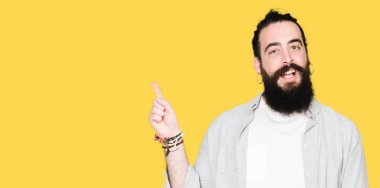 Uzun saçlı, sakallı ve küpeli genç adam yüzünde kocaman bir gülümseme ile, kameraya bakarak yan el ve parmak ile işaret.