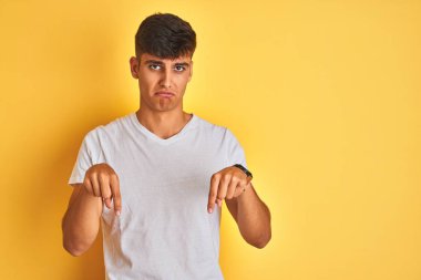 İzole sarı arka plan üzerinde duran beyaz t-shirt giyen genç hintli adam üzgün ve üzgün görünümlü, parmakları ile yön gösteren, mutsuz ve depresif.