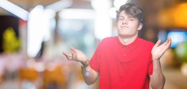 Üzerinde kırmızı t-shirt giyen genç yakışıklı adam arka plan clueless ve şaşkın bir ifadeyle kollar ve eller kaldırdı izole. Şüphe kavramı.