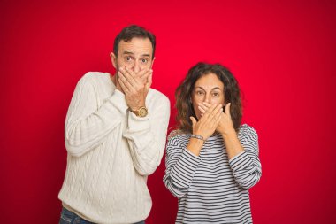 Izole kırmızı arka plan üzerinde kış kazak giyen güzel orta yaş çift hata için elleri ile ağız kapsayan şok. Gizli kavram.