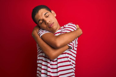 Genç yakışıklı Arap adam izole kırmızı arka plan üzerinde çizgili t-shirt giyen kendini mutlu ve olumlu sarılma, kendinden emin gülümseyen. Öz sevgi ve öz bakım