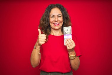 Orta yaş yaşlı kadın büyük bir gülümseme ok işareti yapıyor mutlu kırmızı izole arka plan üzerinde dolar demet tutan, parmaklarla başparmak, mükemmel işareti