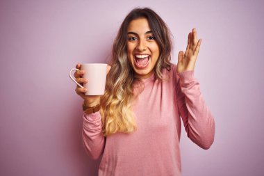 Genç güzel kadın pembe izole arka plan üzerinde kahve bir fincan tutan çok mutlu ve heyecanlı, kazanan ifade büyük bir gülümseme ile çığlık zafer kutluyor ve ellerini kaldırdı