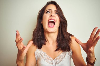 Genç güzel kadın beyaz izole arka plan üzerinde duran t-shirt giyen deli ve deli bağırarak ve agresif ifade ve kollar kaldırdı ile bağırıyor. Hayal kırıklığı kavramı.