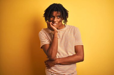 Çizgili t-shirt giyen dreadlocks ile Afro adam izole sarı arka plan üzerinde duran stresli ve ağız tırnak ısırma elleri ile sinir görünümlü. Anksiyete sorunu.