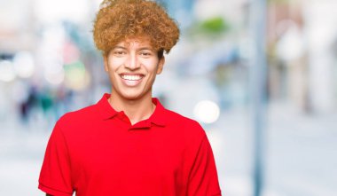 Afro saçlı genç yakışıklı adam yüzünde mutlu ve serin bir gülümseme ile kırmızı t-shirt giyiyor. Şanslı kişi.