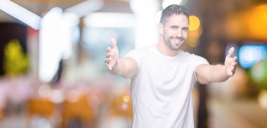 Sarılmak için kollarını açarak gülümseyerek kameraya bakarak izole arka plan üzerinde rahat beyaz t-shirt giyen genç adam. Neşeli ifade kucaklayan mutluluk.