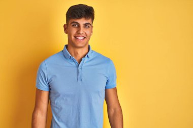 Mavi polo giyen genç hintli adam yüzünde mutlu ve serin bir gülümseme ile izole sarı arka plan üzerinde duruyor. Şanslı kişi.