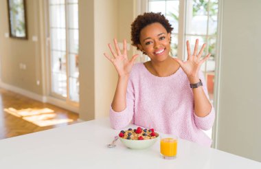 Genç afro-amerikan kadın evde sabah sağlıklı kahvaltı yapıyor gösteren ve parmakları ile işaret ederken emin ve mutlu gülümseyerek on numara.