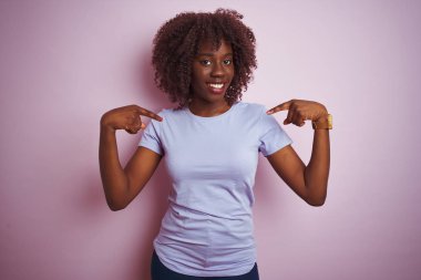 Genç afrikalı afro kadın izole pembe arka plan üzerinde duran t-shirt giyen yüzünde gülümseme ile emin görünümlü, parmakları gururlu ve mutlu kendini işaret.