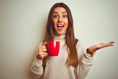 Genç güzel kadın izole beyaz arka plan üzerinde duran kahve kırmızı fincan içme çok mutlu ve heyecanlı, kazanan ifade büyük bir gülümseme ile çığlık zafer kutluyor ve ellerini kaldırdı