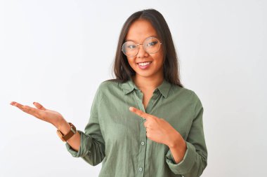 İzole beyaz arka plan üzerinde yeşil gömlek ve gözlük giyen genç Çinli kadın şaşırttı ve el ile sunarken kameraya gülümseyerek ve parmak ile işaret.