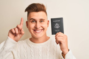 İzole beyaz arka plan üzerinde ABD pasaportu tutan genç yakışıklı adam mutlu yüz, bir numaralı ile bir fikir ya da soru işaret parmağı ile şaşırttı