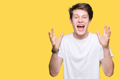 Üzerinde rahat beyaz t-shirt giyen genç yakışıklı adam deli kutluyor arka plan izole ve başarı için kaldırdı kollarını açarak hayrete ve heyecanlı çığlık gözleri açık. Kazanan kavramı