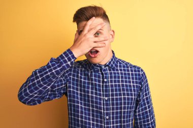 Izole sarı arka plan üzerinde duran rahat gömlek giyen genç yakışıklı adam şok el ile yüz ve gözleri kapsayan gözetleme, utanmış ifade ile parmaklarından bakarak.