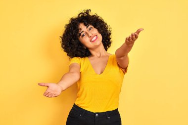 Kıvırcık saçlı genç arap kadın izole sarı arka plan üzerinde duran t-shirt giyen sarılmak için açık kollarla gülümseyen kameraya bakıyor. Mutluluğu kucaklayan neşeli ifade.