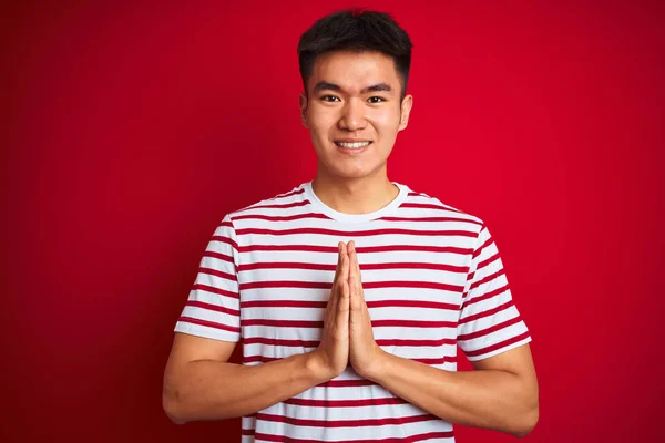 孤立した赤い背景の上に立つストライプのTシャツを着た若いアジア人の男性が 自信を持って笑顔で許しを求めて手をつないで祈る — ストック写真