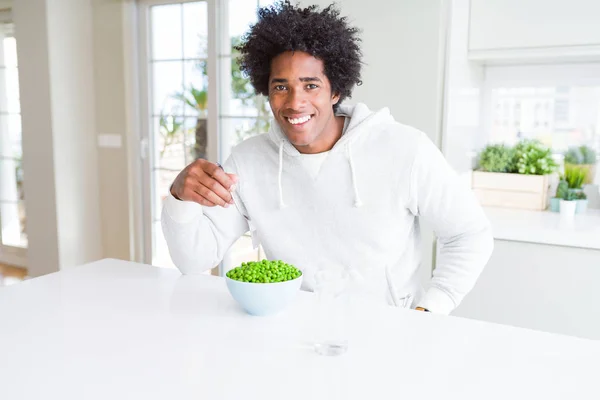 African American man eating fresh green peas at home with a happy face standing and smiling with a confident smile showing teeth