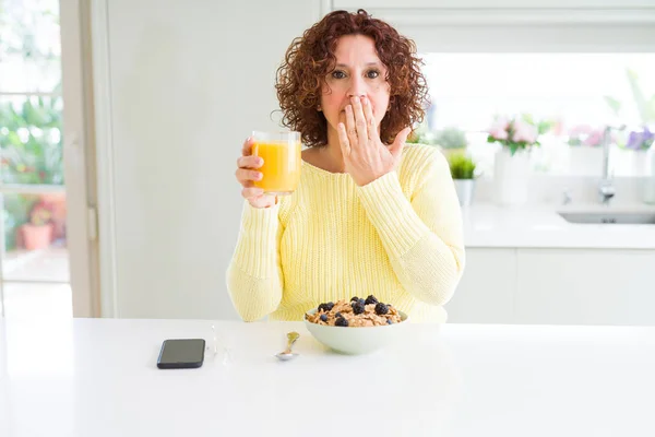 Senior woman eating healthy breakfast in the morning at home cover mouth with hand shocked with shame for mistake, expression of fear, scared in silence, secret concept