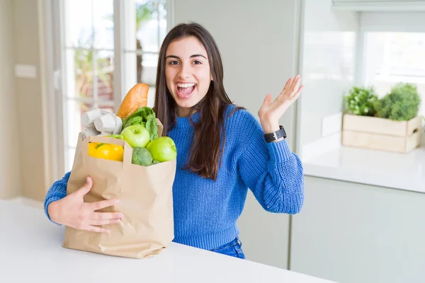 健康的な食料品でいっぱいの紙袋を持つ美しい若い女性は非常に幸せで興奮し 大きな笑顔で叫んで勝利を祝う勝者の表情と手を上げた — ストック写真