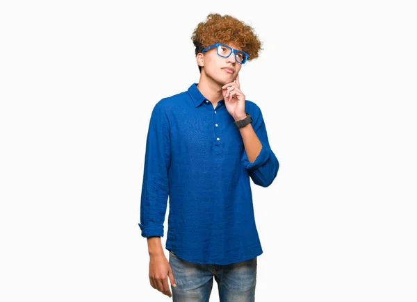 アフロヘアの若いハンサムな男は 質問を考えてあごに手を当てた青いメガネをかけた ペンシブな表情 思慮深い顔で微笑む 疑いの概念 — ストック写真