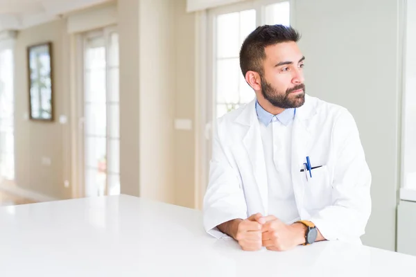 Handsome hispanic doctor or therapist man wearing medical coat at the clinic looking to side, relax profile pose with natural face with confident smile.