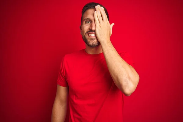 赤い孤立した背景の上にカジュアルなTシャツを着た若いハンサムな男は 手で片目を覆い 顔に自信のある笑顔と驚きの感情 — ストック写真