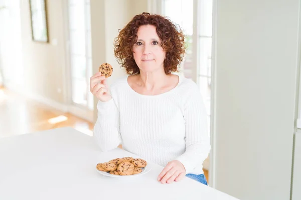 真剣に考えるスマートな顔に自信のある表情で自宅でチョコチップクッキーを食べるシニア女性 — ストック写真