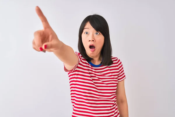 Young beautiful chinese woman wearing red striped t-shirt over isolated white background Pointing with finger surprised ahead, open mouth amazed expression, something on the front
