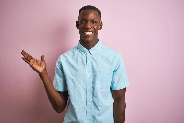 孤立したピンクの背景の上に立つ青いカジュアルなシャツを着たアフリカ系アメリカ人男性は カメラを見て手のひらを指差して陽気な提示と笑顔を浮かべている — ストック写真