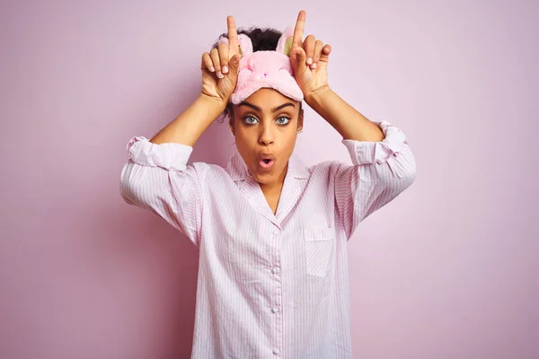 年轻的非洲裔美国妇女穿着睡衣和面具在孤立的粉红色背景做有趣的手势用手指在头上作为牛角 — 图库照片