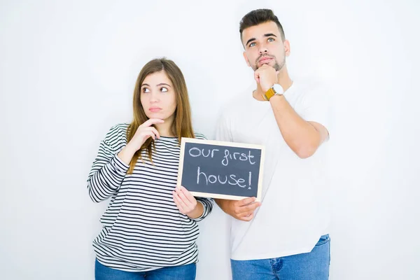 Young couple holding blackboard with our first home text over white isolated background serious face thinking about question, very confused idea