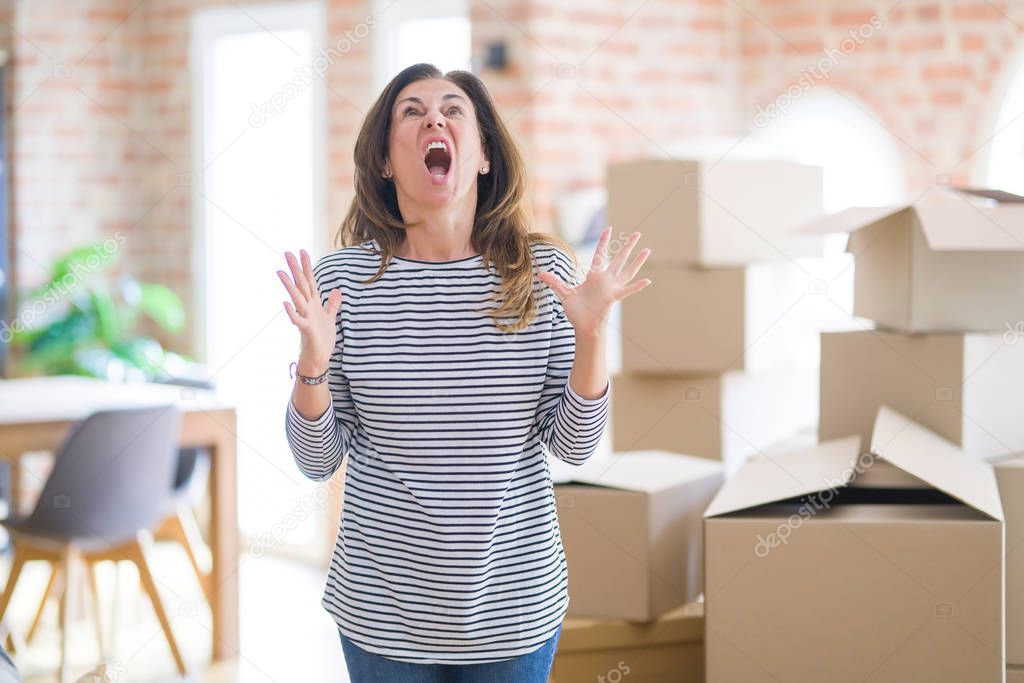 Middle age woman moving to a new house arround cardboard boxes crazy and mad shouting and yelling with aggressive expression and arms raised. Frustration concept.