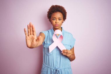 Genç afro-amerikan kadın ciddi ve kendinden emin ifade, savunma jesti ile dur işareti yapıyor açık el ile izole pembe arka plan üzerinde brest kanser kurdele tutan