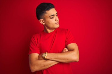 Izole kırmızı arka plan üzerinde duran t-shirt giyen genç Brezilyalı adam kolları ile yan bakarak ikna ve kendinden emin geçti