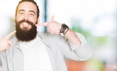 Uzun saç, sakal ve kendinden emin gösterilen ve işaret parmakları diş ve ağız ile gülümseyen küpe olan genç adam. Sağlık kavramı.