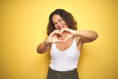 Kıvırcık saçlı orta yaş yaşlı kadın sarı izole arka plan üzerinde duran aşk kalp sembolü ve elleri ile şekil gösteren gülümseyerek. Romantik kavram.