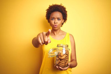 Sarı arka plan üzerinde çikolata cipsi kurabiye kavanoz tutan Afrikalı Amerikalı kadın kameraya parmak ile işaret ve size, el işareti, önden olumlu ve kendinden emin jest