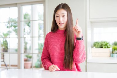 Güzel Asyalı kadın başarılı bir fikir ile parmak işaret beyaz masada pembe kazak giyiyor. Çıktı ve mutlu. Bir numara.